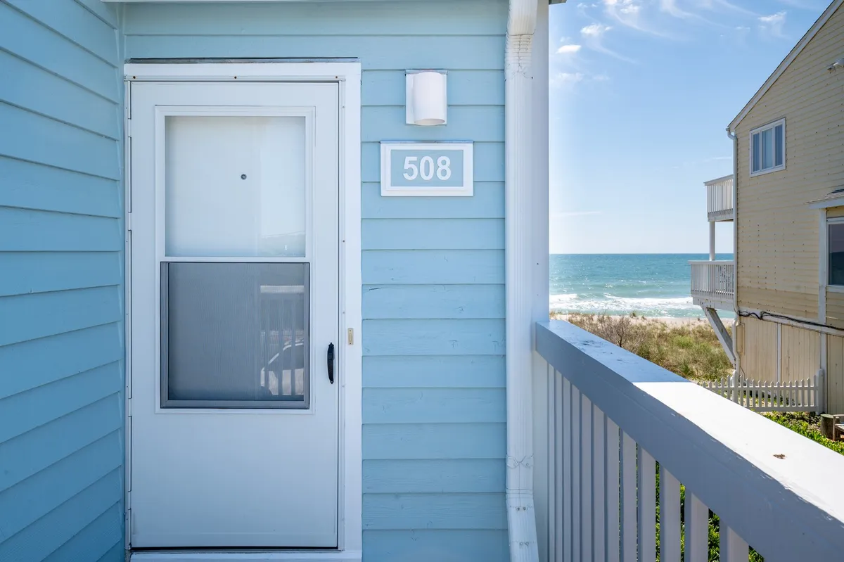 A welcoming entrance with a clear ocean view, the front door framed by coastal landscaping and the distant blue water. The porch area invites guests to step outside and enjoy the natural beauty and sea breeze.