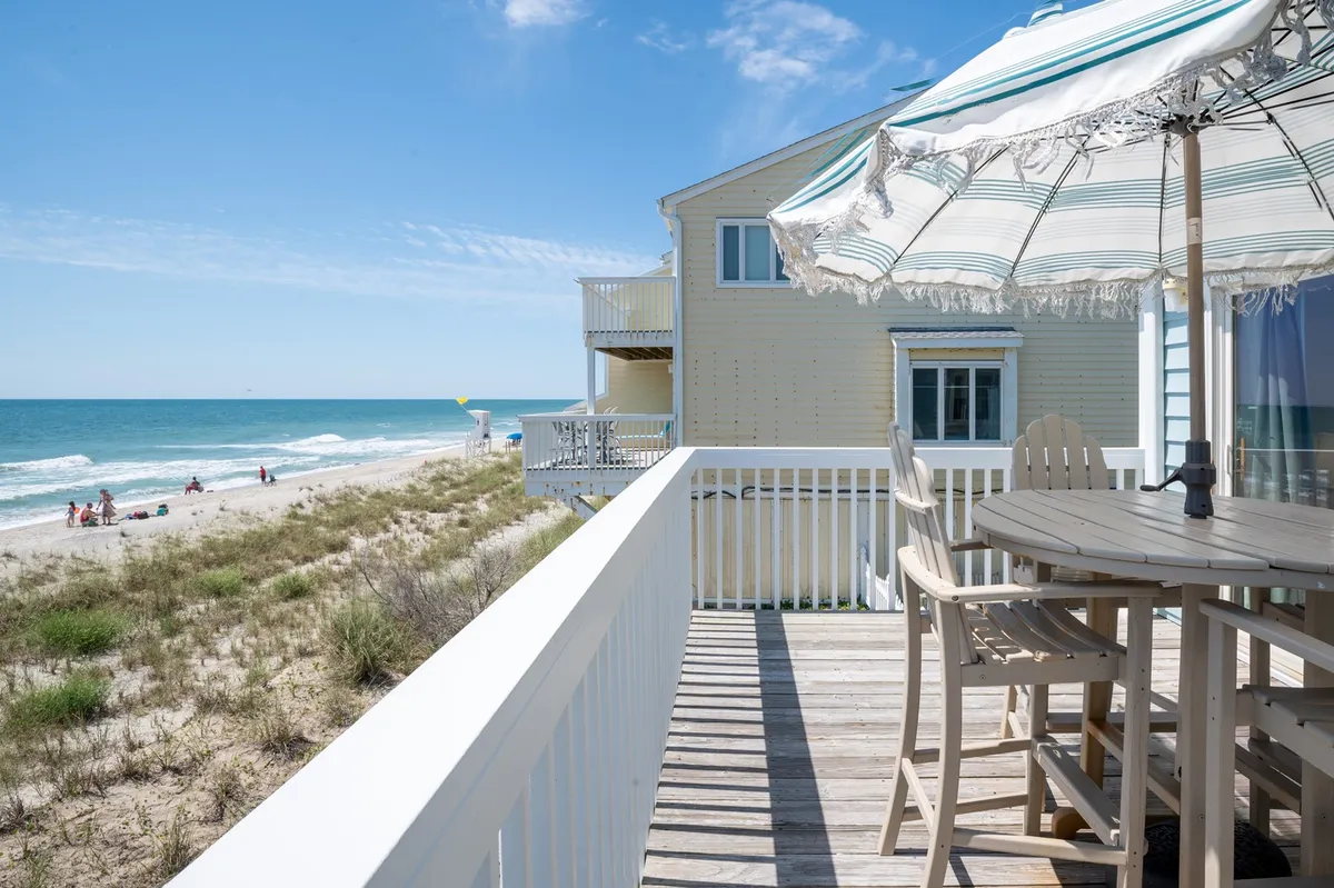 A beachfront deck furnished with comfortable seating and a beach umbrella, overlooking the ocean. The sandy beach stretches just beyond, inviting guests to relax, stroll, or enjoy meals outdoors. Early morning light hints at the possibility of sunrise coffee and tranquil moments by the shore.