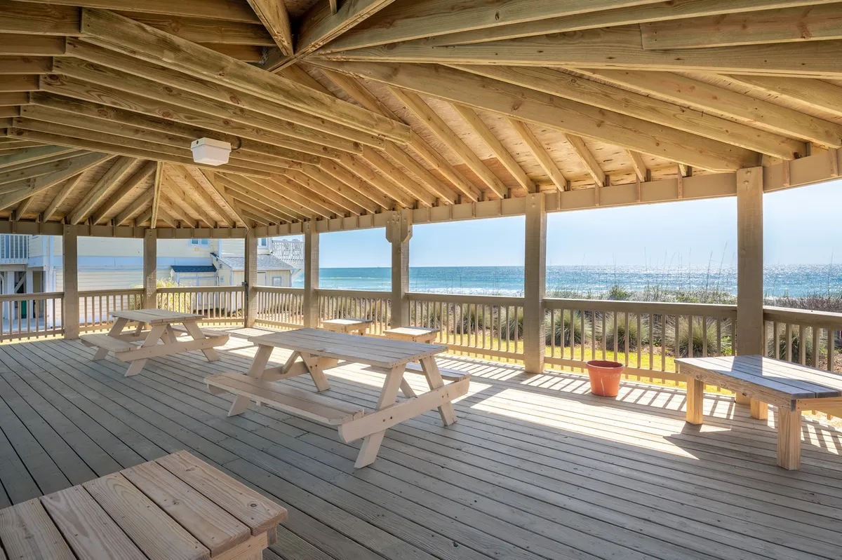 A cheerful outdoor area with sturdy wooden picnic tables and expansive sea views. Wooden beams frame the scene, creating a welcoming space for casual dining, sunset watching, or relaxed gatherings by the ocean.