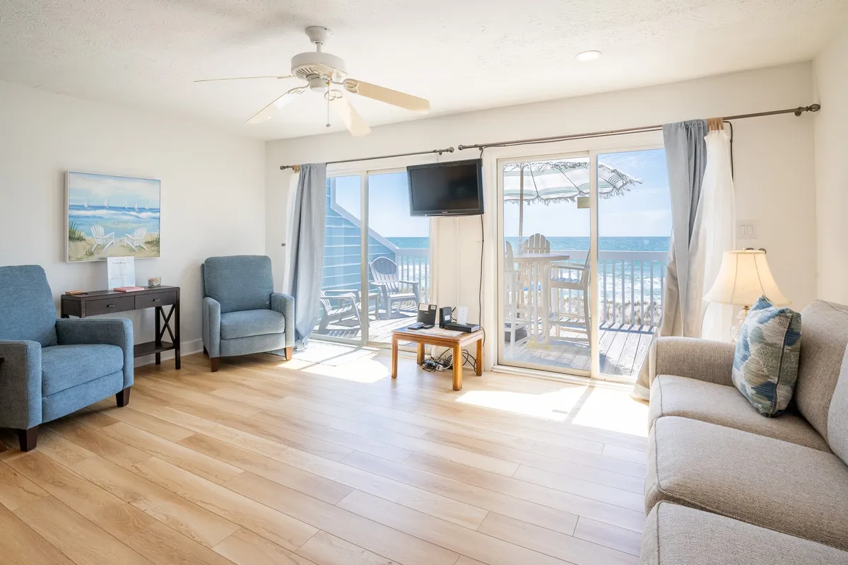 A bright living room features comfortable seating arranged near a large sliding glass door, with inviting beach-themed artwork and a glimpse of the ocean beyond. The space feels airy and tranquil, filled with natural light and views of the shoreline.