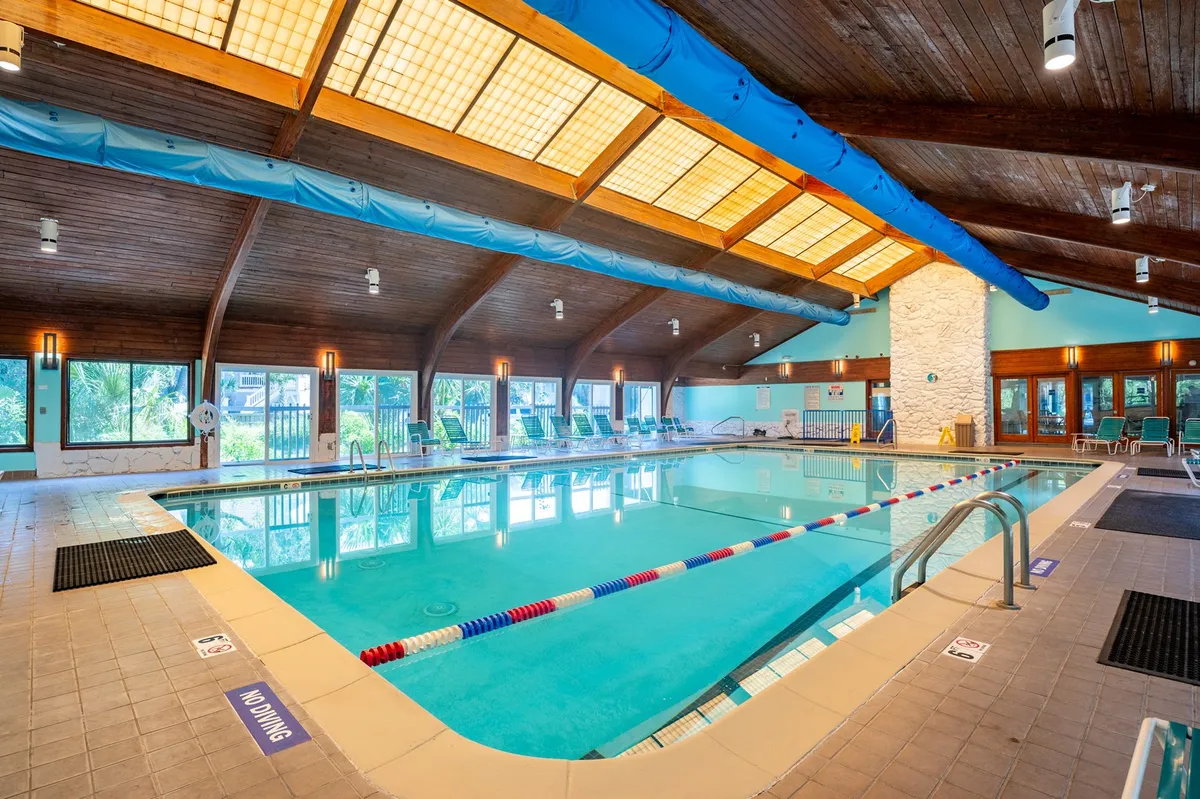 A bright, inviting indoor pool area with a wooden ceiling, expansive windows flooding the space with natural light, and plush poolside lounge chairs. People can be seen swimming, relaxing by the water, and gathering in small groups, suggesting both activity and relaxation.