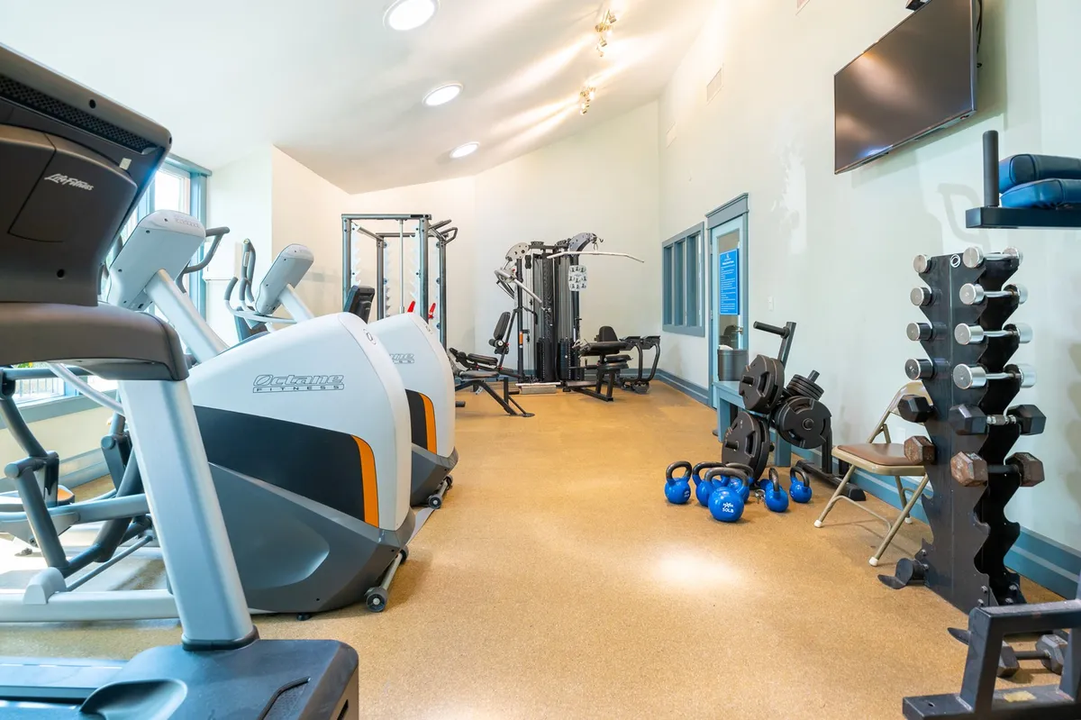 A modern exercise room filled with treadmills, ellipticals, weight machines, and free weights. The energetic scene shows several people working out, some attending group fitness classes, fostering a sense of wellness and activity.