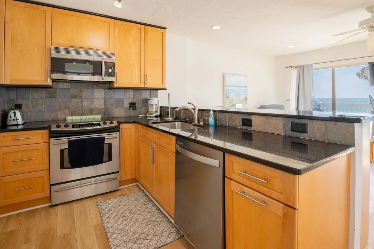 A modern kitchen with stainless steel appliances, granite countertops, and warm wood cabinetry. A large window frames a picturesque ocean view, infusing the space with natural light and connecting the kitchen to the scenic surroundings.