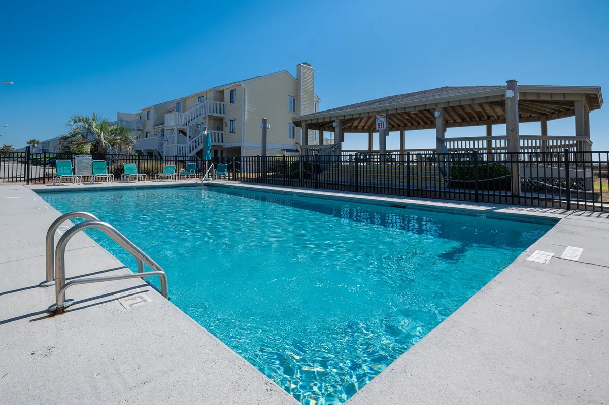 A large swimming pool area surrounded by lounge chairs and a gazebo, with resort buildings in the background. The pool is a hub for both relaxation and socializing, with ample space for groups or quiet reflection.