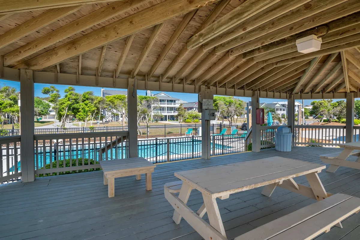 An exterior scene featuring inviting wooden picnic tables alongside a sparkling swimming pool, with children and families visible enjoying the water and the outdoor space. Greenery surrounds the area, and a gazebo offers a shaded spot for gatherings.