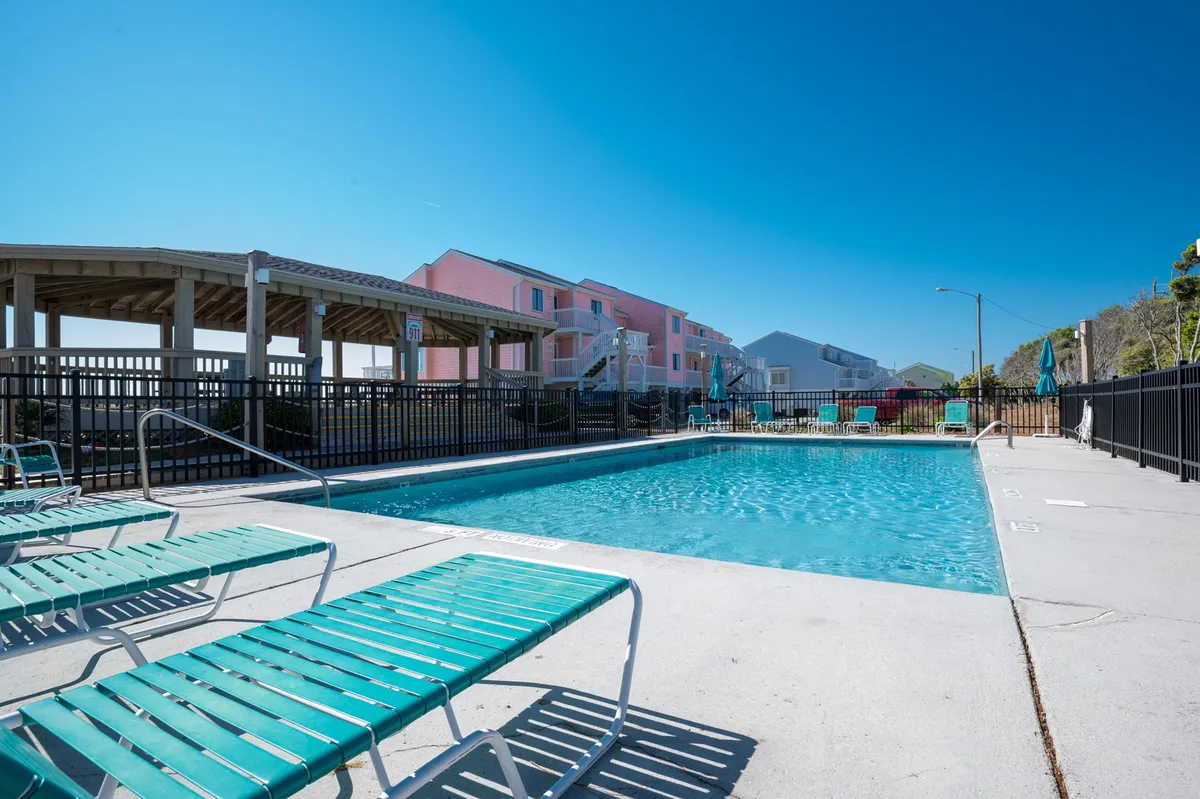 An outdoor swimming pool surrounded by comfortable lounge chairs and adjacent buildings. The scene is set during golden hour, with a relaxed group enjoying the water and others sunbathing on deck chairs, capturing the essence of leisure and social moments.