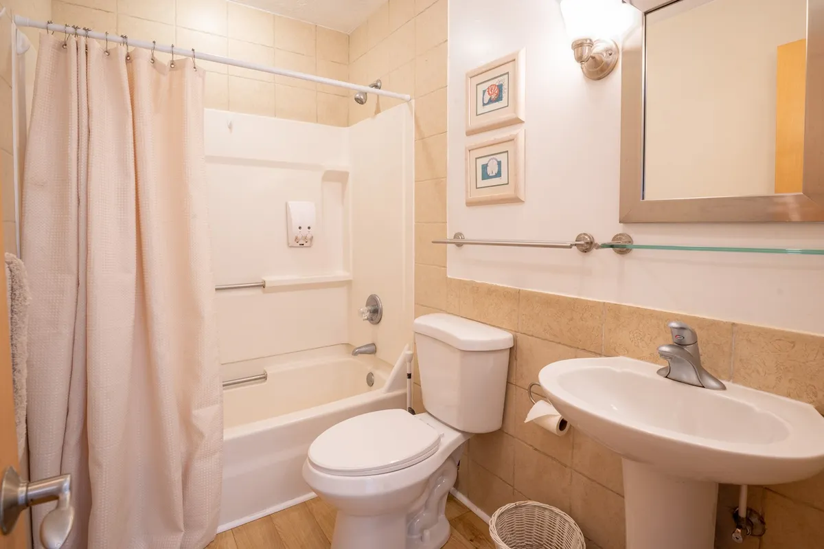 A bright bathroom scene featuring a shower area, toilet, and sink, with decorative frames on the walls and gentle morning light streaming in. The space feels fresh, private, and inviting—ideal for quick rinses or relaxing soaks.