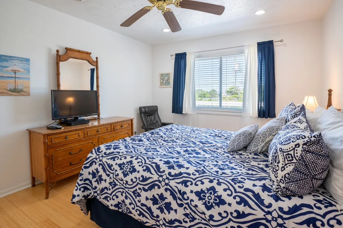 A cozy bedroom with large patterned bedding, a wooden dresser, a television, and a window with curtains allowing natural light inside. The room feels inviting and perfect for both relaxation and privacy.