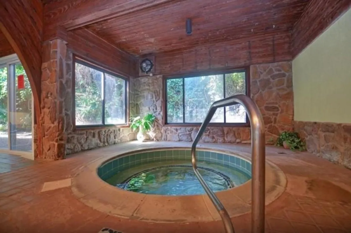 A serene hot tub area with stone walls, large windows that let in abundant natural light, and a touch of greenery. The spa-like ambiance is complemented by people relaxing in the hot tub and enjoying tranquil views outdoors.